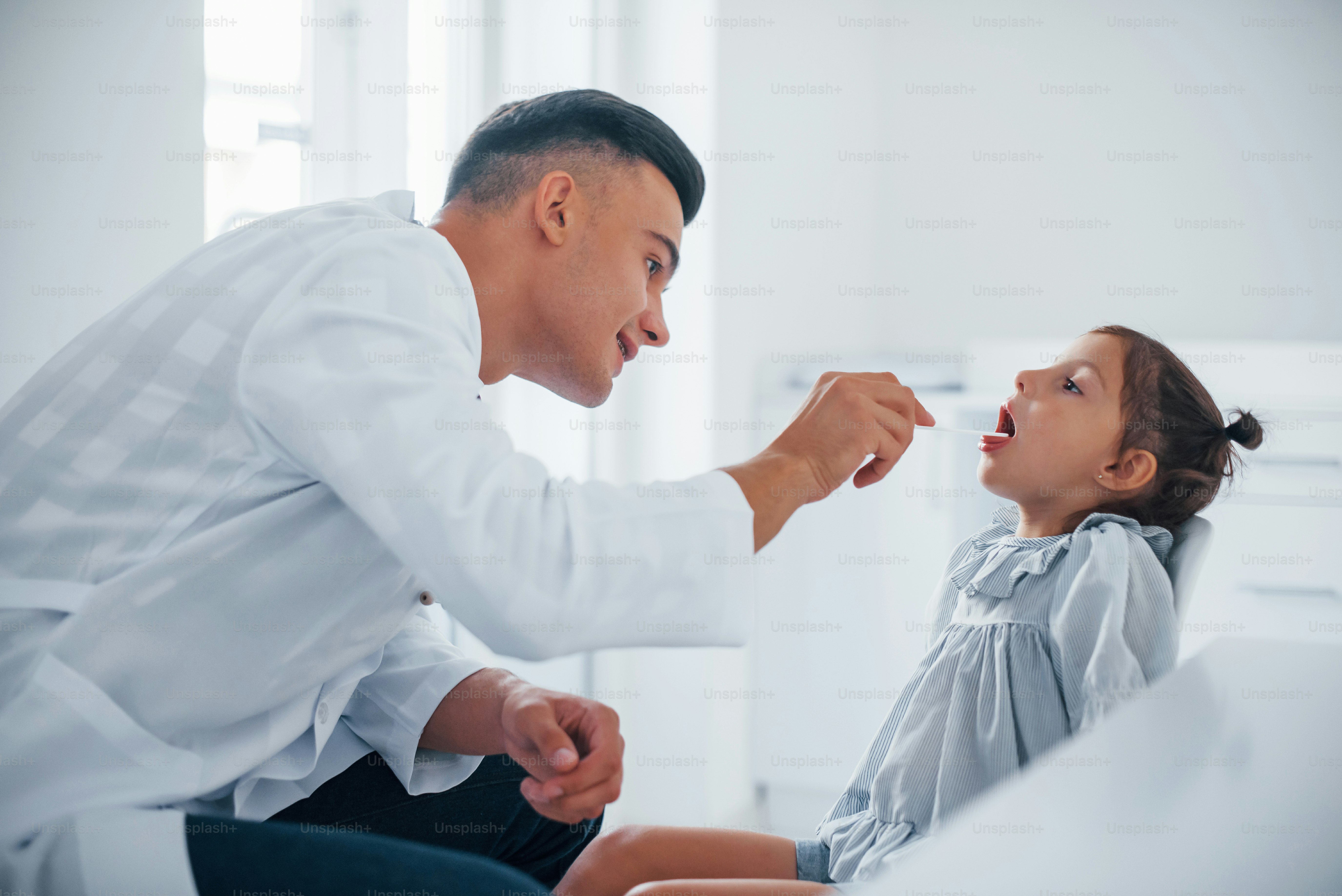 Uses tonsil to check throat. Young pediatrician works with little female visitor in the clinic.